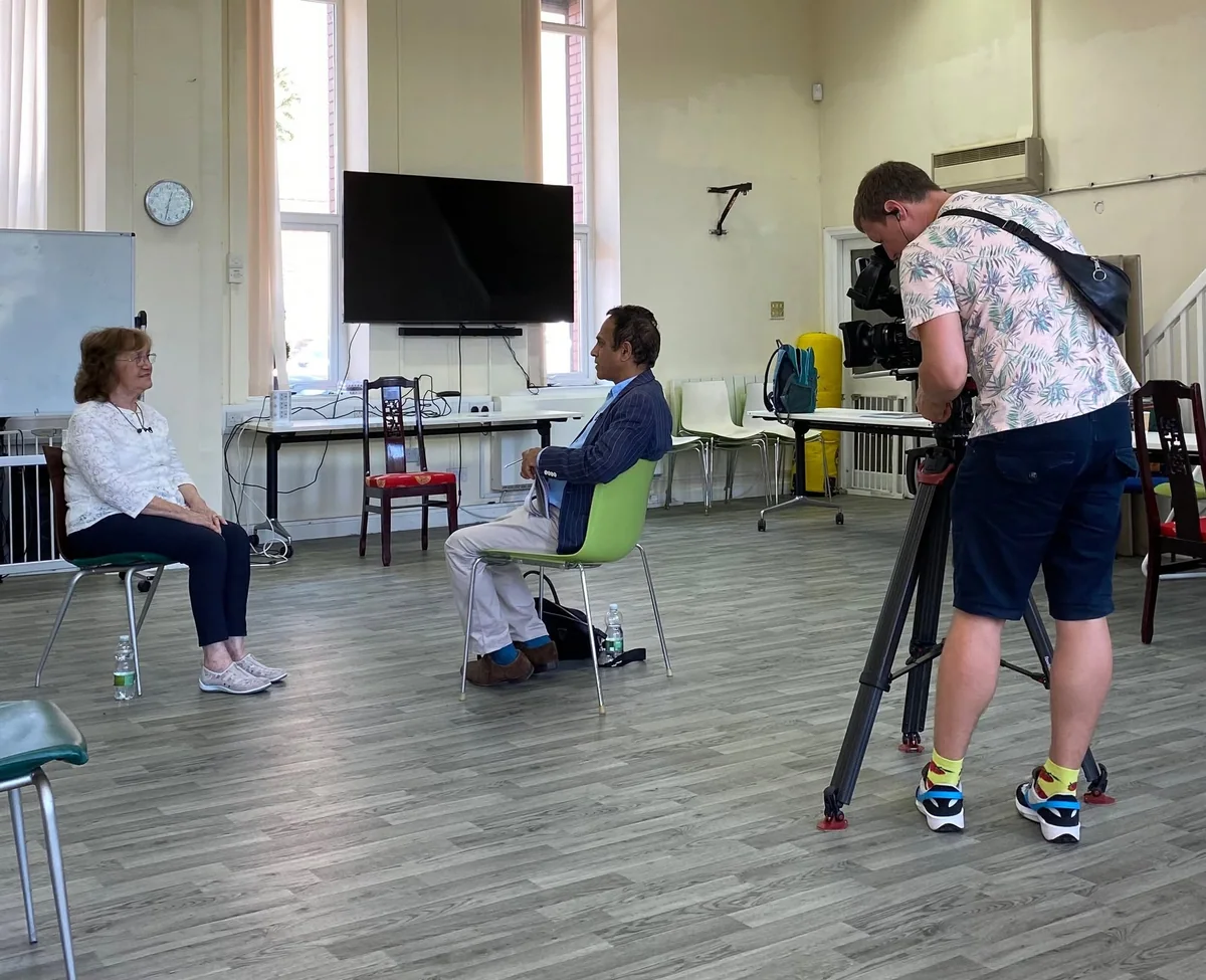 A woman speaking with an interviewer while a camera operator records the conversation.