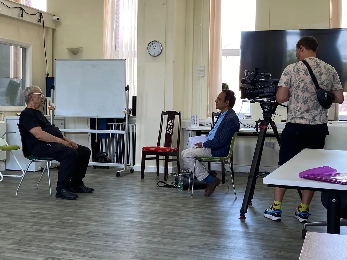 A seated man in conversation with an interviewer and a camera operator.
