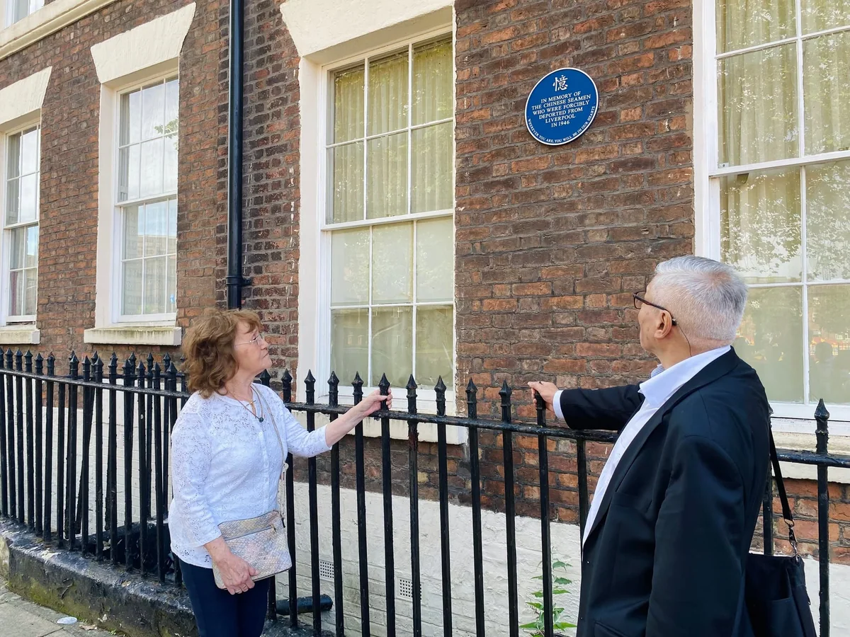 Two people talking beside the blue memorial plaque in Liverpool.